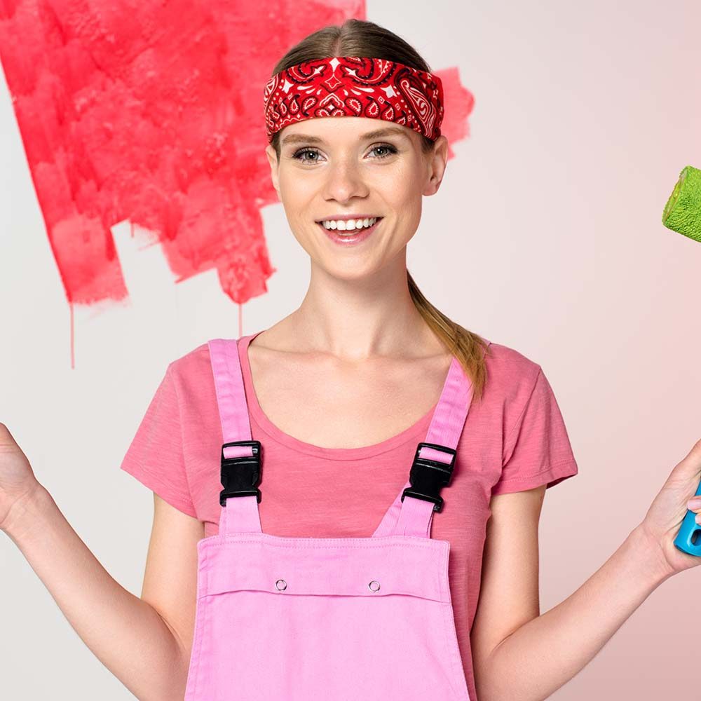 smiling woman in working overall holding paint rollers in front of painted wall